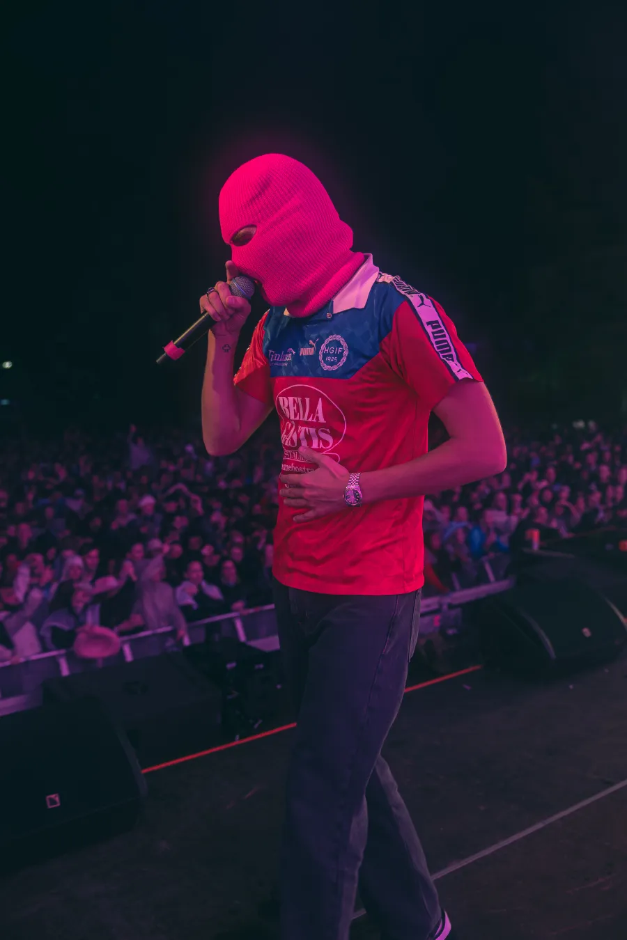 An artist in a pink hoodie singing into a microphone, with the audience in the background