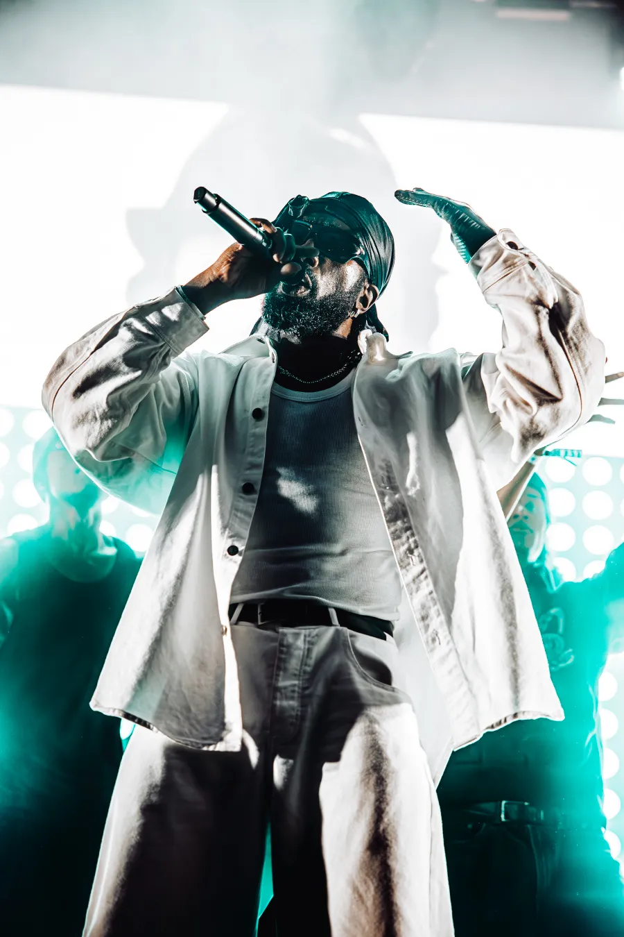 A male artist in a white outfit singing into a microphone with one hand raised.