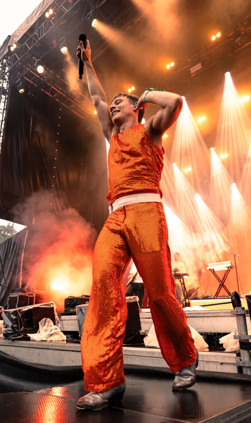 An artist in an orange outfit standing on stage with raised hands